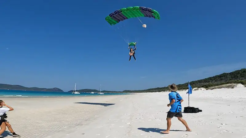 Experience a thrilling tandem skydive at Whitehaven Beach: parachutist landing, onlookers, sailing boats, lush hills, video and photos.