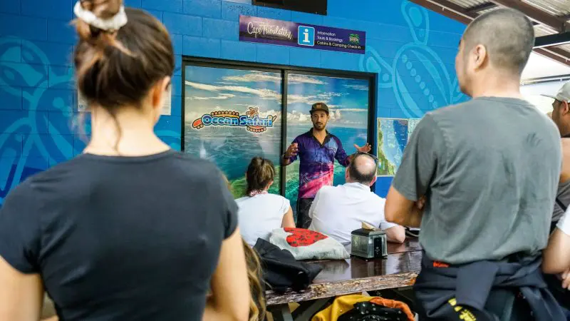 An expert guide presents the Great Barrier Reef Eco Adventure to an attentive group seated indoors by a vibrant ocean-themed blue wall.