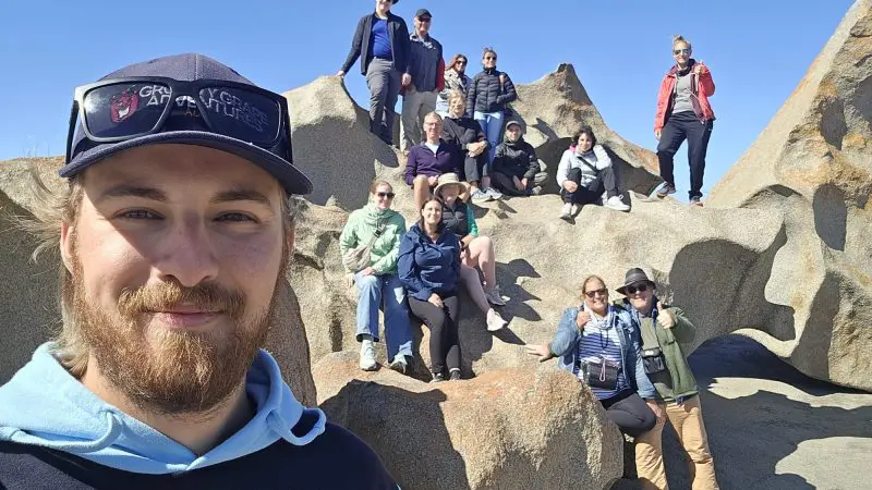 A smiling group on rugged rocks beneath a clear sky captures a selfie during their Kangaroo Island 2 Day Wildlife Adventure tour.