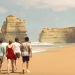 Group of four enjoying a scenic walk on sandy beach below dramatic cliffs and crashing waves during 1 Day Great Ocean Road Eco Tour.