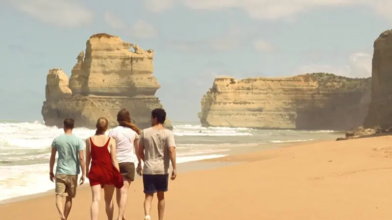 Group of four enjoying a scenic walk on sandy beach below dramatic cliffs and crashing waves during 1 Day Great Ocean Road Eco Tour.