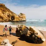 Couple walks hand in hand on a sandy beach during a 1 Day Great Ocean Road Rainforest Tour, with dramatic cliffs and rocky boulders.