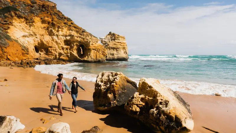 Couple strolls hand in hand on sandy beach at sunrise during 1 Day Great Ocean Road Reverse Eco Tour, enjoying scenic coastal views.