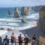 Travellers on a 1 Day Great Ocean Road Rainforest Tour admire the iconic Twelve Apostles by the picturesque, partly cloudy coastline.
