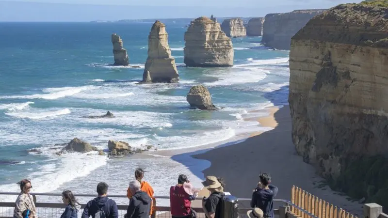 Travellers on a 1 Day Great Ocean Road Rainforest Tour admire the iconic Twelve Apostles by the picturesque, partly cloudy coastline.