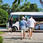 Three guests approach a Peninsula Hot Springs Spa Shuttle Bus under clear blue skies, surrounded by lush trees and natural scenery.