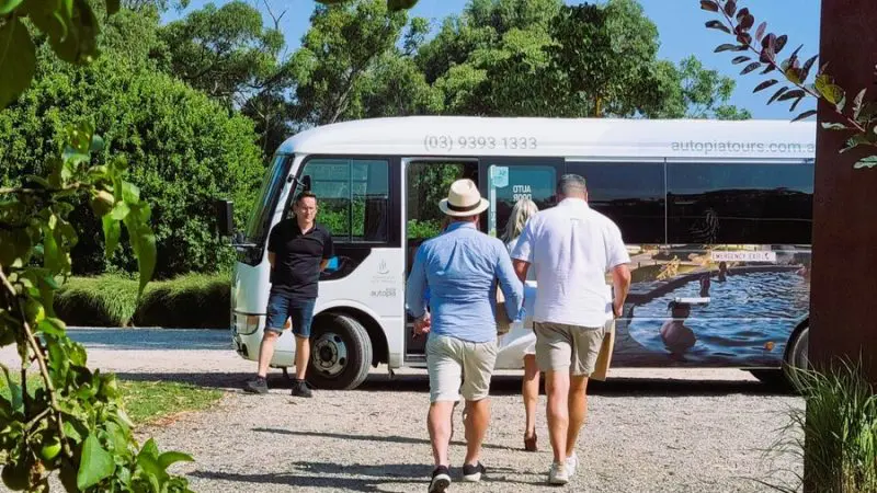 Three guests approach a Peninsula Hot Springs Spa Shuttle Bus under clear blue skies, surrounded by lush trees and natural scenery.
