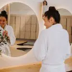 Smiling woman in a white dressing gown admires her reflection during a relaxing Peninsula Hot Springs Spa day, enjoying shuttle bus entry.