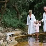 Happy family of two adults and a child in spa robes walking barefoot by lush greenery after Peninsula Hot Springs day spa shuttle experience.