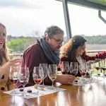 Three friends wine tasting outdoors, laughing on a 1 Day Hunter Valley Scenic Wine Tour amid vineyards under clear skies.