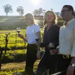Three people walk through lush Hunter Valley vineyards on a one-day wine tour, smiling and holding vibrant red wine glasses in hand.