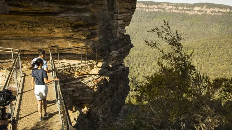 Experience breathtaking Blue Mountains views as two adventurers walk a fenced cliffside trail on a Sunset Wilderness Tour.