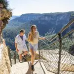 Smiling couple hiking up a rocky trail on a 1 Day Blue Mountains Sunset Wilderness Tour, scenic panoramic mountain views in the background.