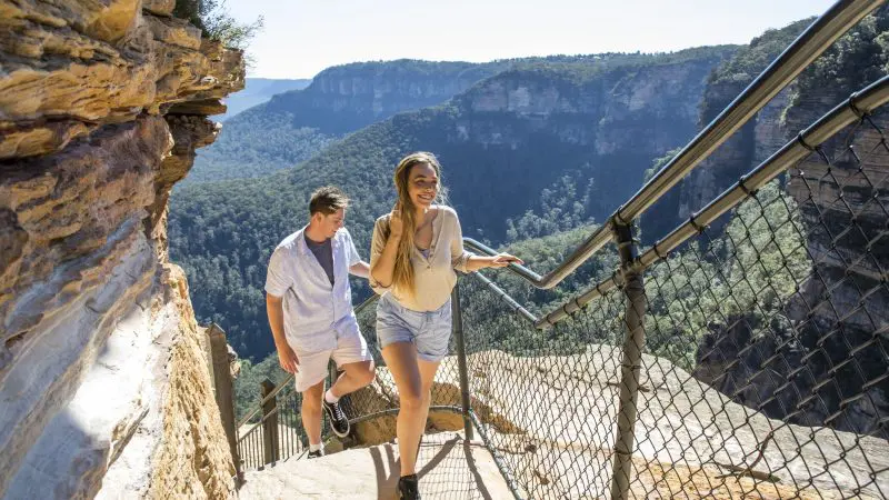 Smiling couple hiking up a rocky trail on a 1 Day Blue Mountains Sunset Wilderness Tour, scenic panoramic mountain views in the background.