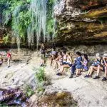 Adventurers on a rocky ledge take in lush Blue Mountains views as two climbers ascend nearby during a 1 Day Sunset Wilderness Tour.