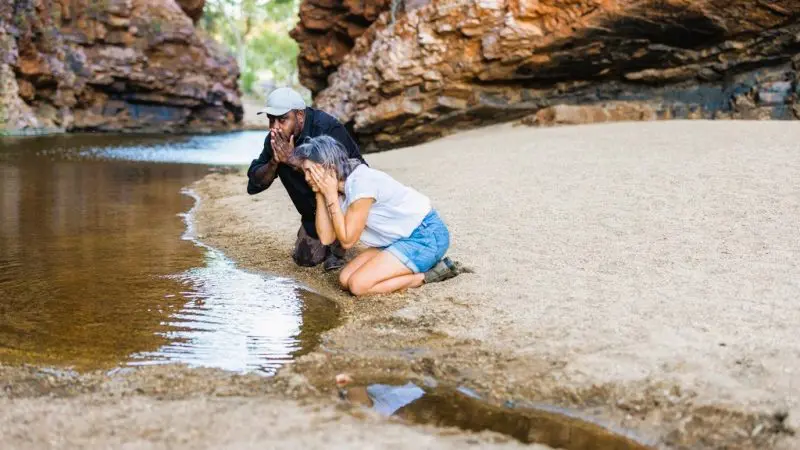 Two adventurers kneel by a pristine creek in the West MacDonnell Ranges, splashing faces on a 1 Day Standley Chasm guided tour.