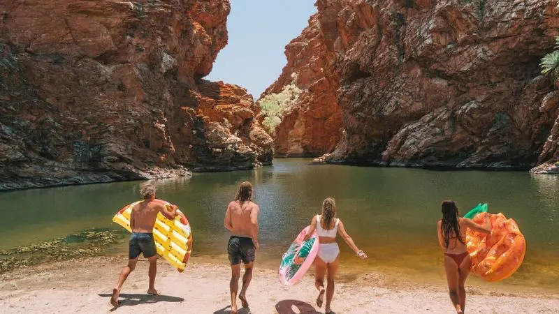 Four friends with colourful pool floats head to a scenic waterhole in the West MacDonnell Ranges near iconic Standley Chasm, Australia.