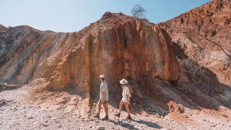 Two adventurers in hats hike along a sunlit cliff on a 1 Day West MacDonnell Ranges and Standley Chasm guided tour, Australia.