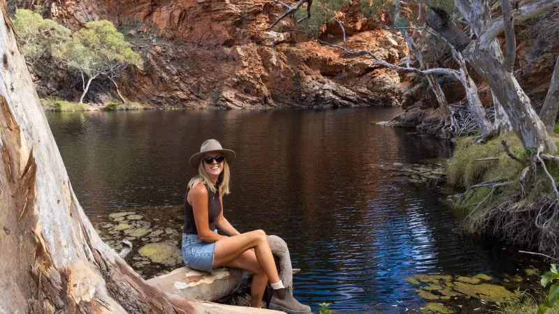 A woman wearing a hat and sunglasses relaxes by a scenic rocky waterhole during a 1-Day West MacDonnell Ranges Standley Chasm tour.