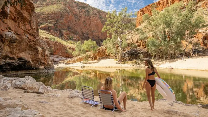Two women relaxing by a riverside under rocky cliffs and trees in West MacDonnell Ranges on a guided Standley Chasm day tour.
