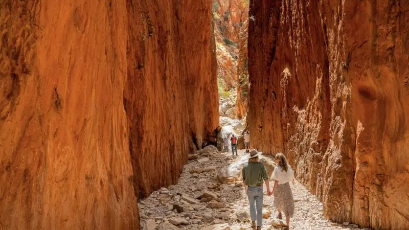 Couples explore Standley Chasm on a 1 Day West MacDonnell Ranges tour, framed by towering red-orange canyon walls and rugged scenery.