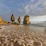 Couple walking along sandy beach at sunset on Great Ocean Road tour from Melbourne, with scenic waves and rocky coastline backdrop.
