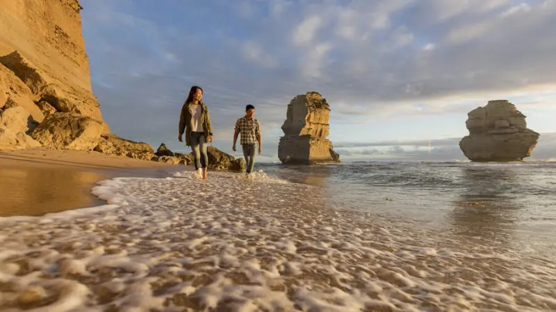 Couple walking along sandy beach at sunset on Great Ocean Road tour from Melbourne, with scenic waves and rocky coastline backdrop.
