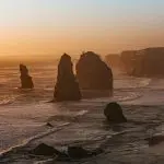 Dramatic rock formations emerge from the sea at sunset on a Great Ocean Road tour from Melbourne, with waves crashing ashore.