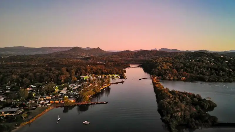 Stunning aerial view of Brunswick River Nature Tour at sunset, featuring boats, riverside homes, lush trees, mountains, and vivid skies.