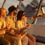 Three friends toast drinks on a BYO Sunset Sparkle Catamaran Cruise on Sydney Harbour, pointing to the iconic Opera House at sunset.