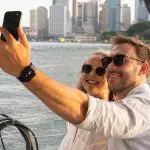 Smiling couple in sunglasses captures a selfie whilst enjoying a Sydney Harbour BYO Sunset Sparkle Catamaran Cruise at sunset.