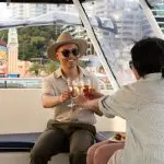 Pair toasts wine glasses aboard a Sydney Harbour catamaran at sunset, BYO cruise, city skyline illuminated in the background.