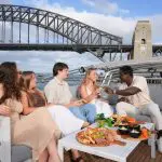 Friends enjoying gourmet food and drinks on a Sydney Harbour Golden Glow Sunset Cruise with iconic Sydney Bridge skyline views.