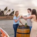 Three friends toast drinks on a Sydney Harbour sunset cruise near the Opera House, smiling and admiring the golden waterfront view.
