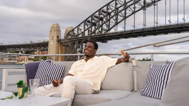 Man relaxing on a luxury yacht with striped cushions, smiling during a Sydney Harbour sunset cruise with iconic bridge views.