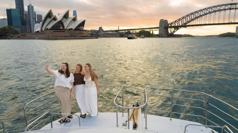 Three women smile for a selfie during a Sydney Harbour sunset cruise, with famous landmarks bathed in golden light behind them.