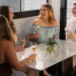 Four women raise a toast with sparkling wine aboard a luxury boat on Sydney Harbour at sunset, golden light reflecting off shimmering waters.