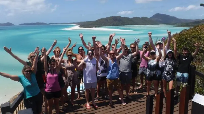 A smiling group enjoys a Thundercat Whitsunday Islands tour on a deck, with stunning turquoise beach and lush mountain views behind.