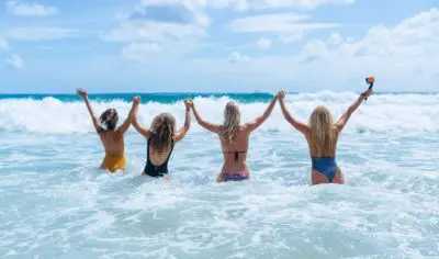 Four women in colourful swimsuits stand in clear sea waves, smiling, holding hands and an action camera during a budget beach holiday.