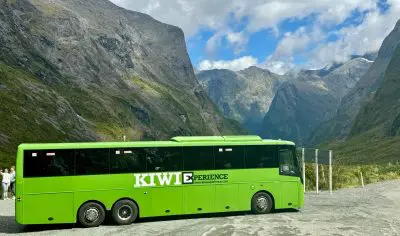 Image of Large green Kiwi Experience bus with the background of Milford sound