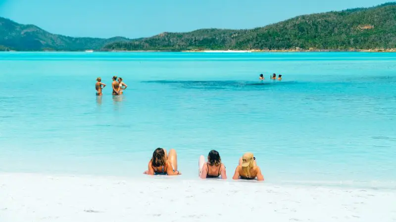 three friends lying on a white sand beach with a group of people standing in the shallows