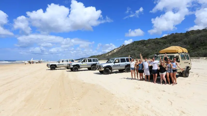 Smiling group of friends posing on a sunny sandy beach near three parked 4x4s under a vibrant blue sky with fluffy clouds.