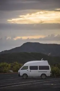White van parked in an empty car park with scenic mountain views and vibrant cloudy sunset skies in the background, perfect for travel.
