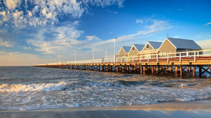 Scenic wooden pier with white railings and charming buildings stretches over tranquil sea beneath a vibrant blue sky with light clouds.