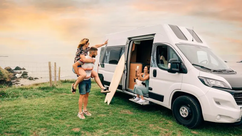 At sunset, two friends by a campervan enjoy a piggyback while two others inside capture holiday photos, creating adventure vibes.