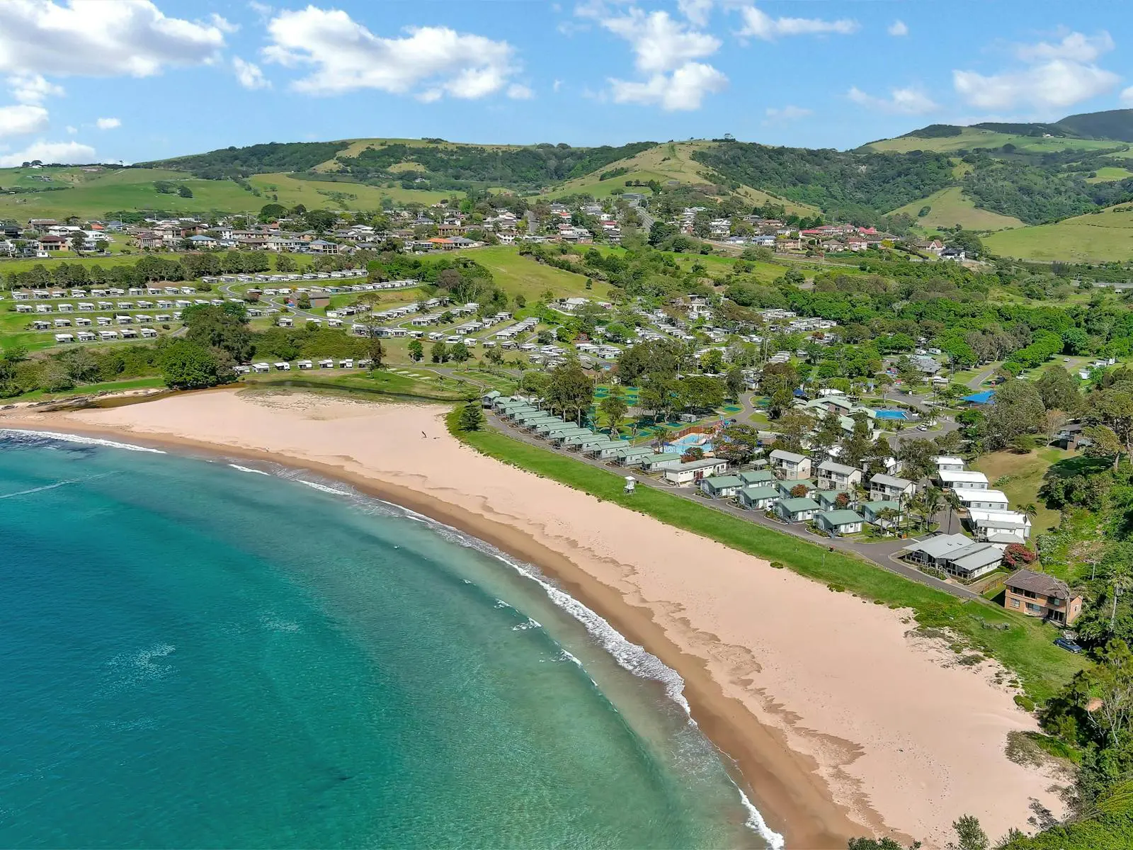 Stunning blue ocean waters meeting soft yellow sand. behind the sand is rolling green hills with a campervan site parked in them