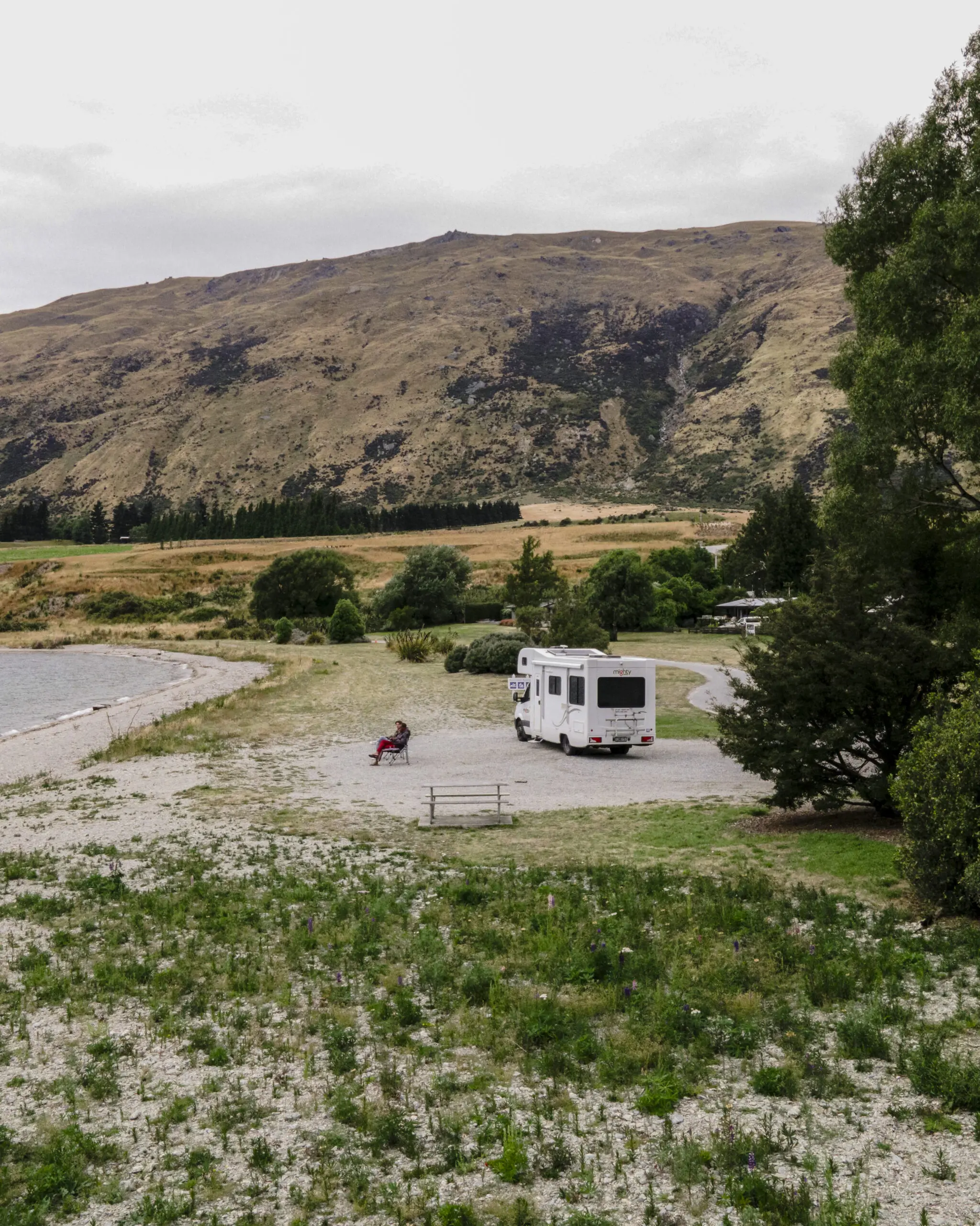 Mighty RV in camping spot by the lake with 2 people sitting on folding chairs outside