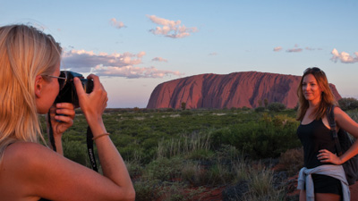 Two women photograph iconic Uluru on a 3-night Rock The Centre Yulara adventure, with dramatic clouds enhancing the Outback landscape.