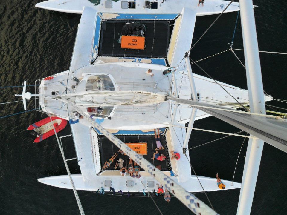 Stunning aerial view of Whitsundays catamaran sailing; people lounging on deck, red dinghy nearby, turquoise water surrounds.