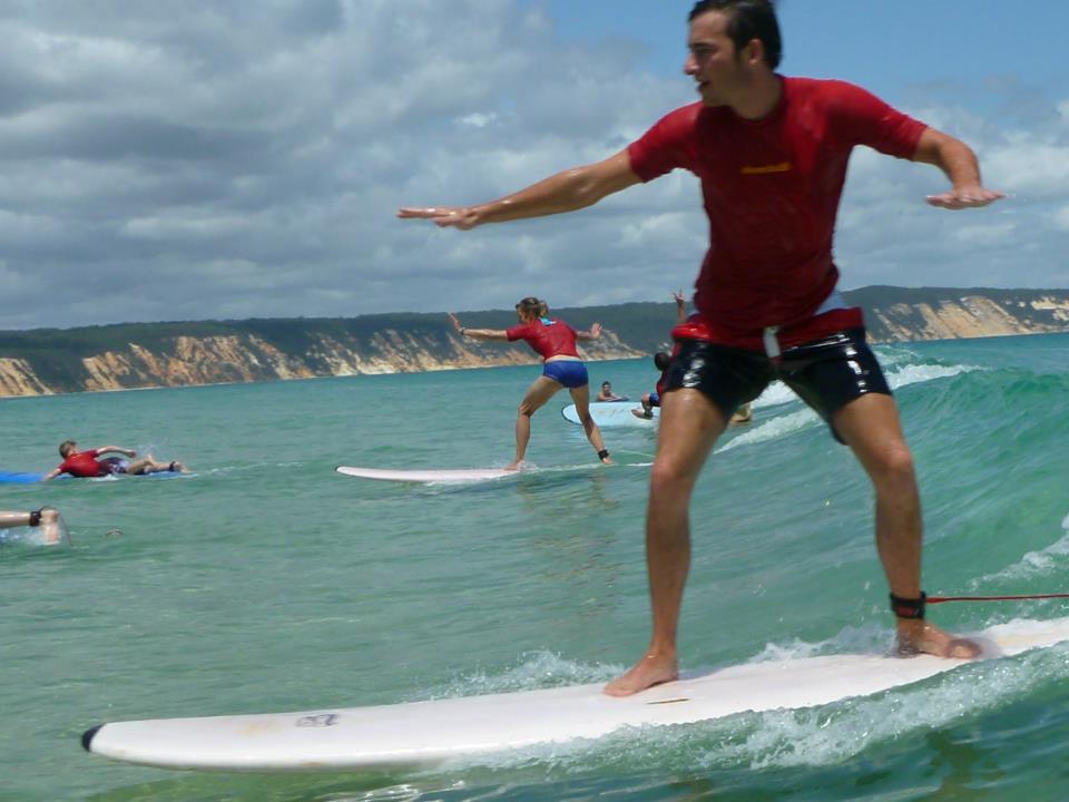 Surfers riding crystal-clear turquoise waves near scenic cliffs, with a Double Island Surfboard Hire 4x4 Adventure vehicle and group nearby.